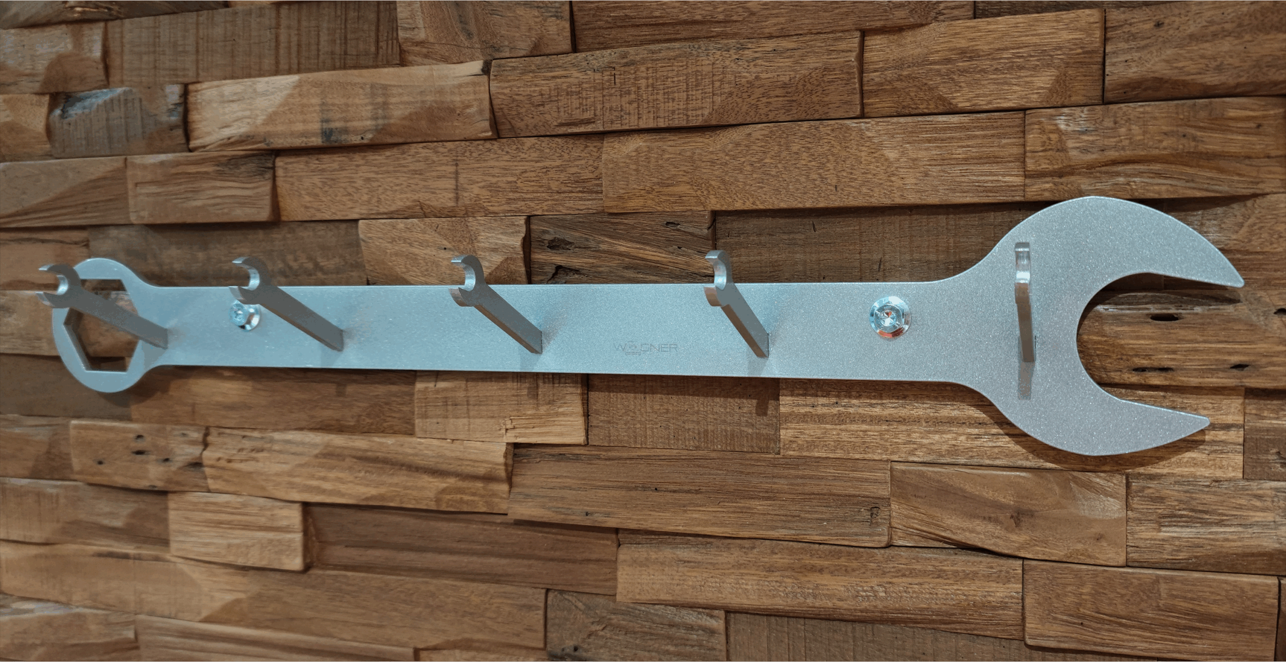 Silberne Wagner-Racing-Wandgarderobe in Form eines großen Schraubenschlüssels mit mehreren schlüsselförmigen Haken vor einer Holzwand.